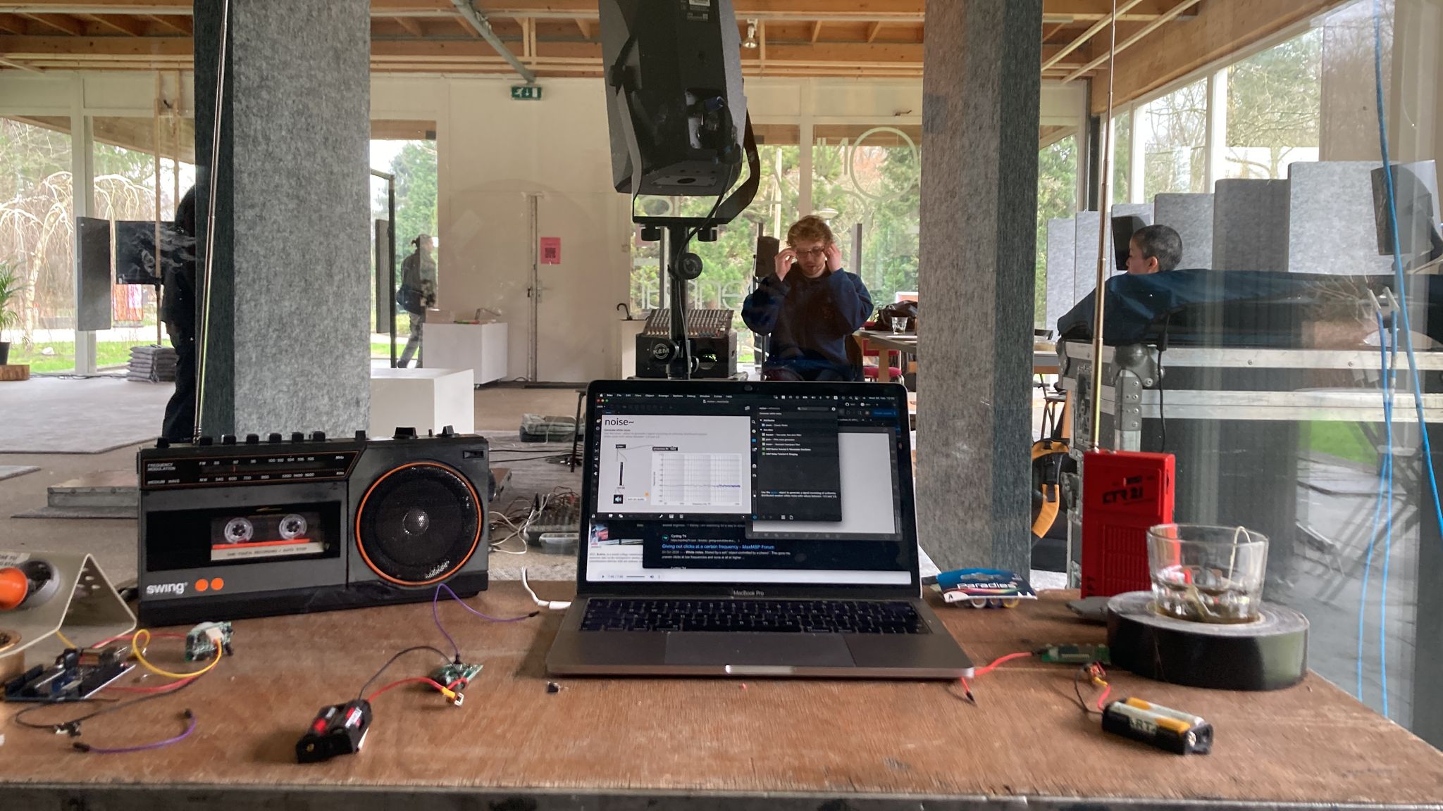 A laptop, two radio and electronic parts scattered on a table facing into a glass house